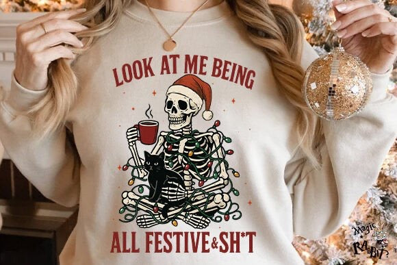 Person wearing a beige sweatshirt with a skeleton and cat design and text 'Look at me being all festive & sh*t'.