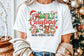 Person wearing a white t-shirt with Christmas-themed text and graphics, standing in front of a decorated Christmas tree.